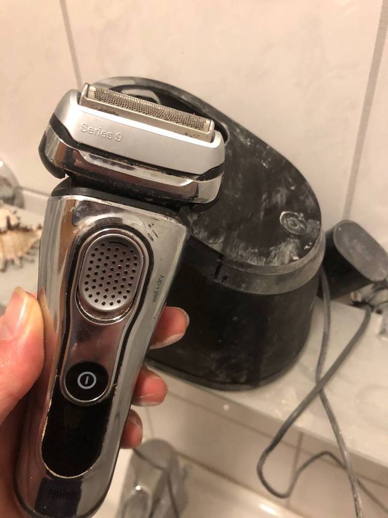 Hand hält einen metallfarbenen Rasierer im Test vor einer schwarzen Ladestation im Badebereich.