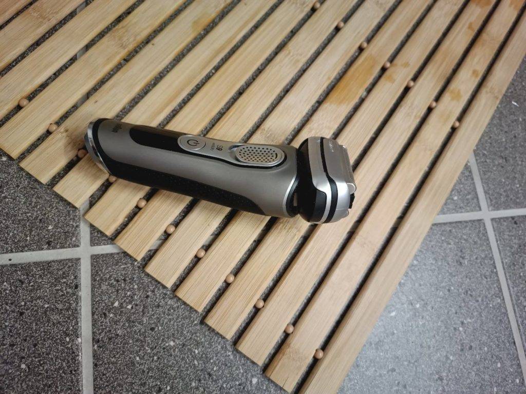 Ein metallfarbener getesteter Rasierer liegt auf der Seite auf einem Badvorleger aus Holz.