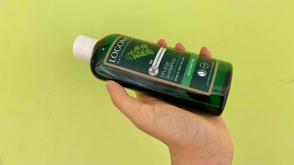 Eine Hand hält eine Flasche Brennnessel-Shampoo im Test vor eine grüne Wand.