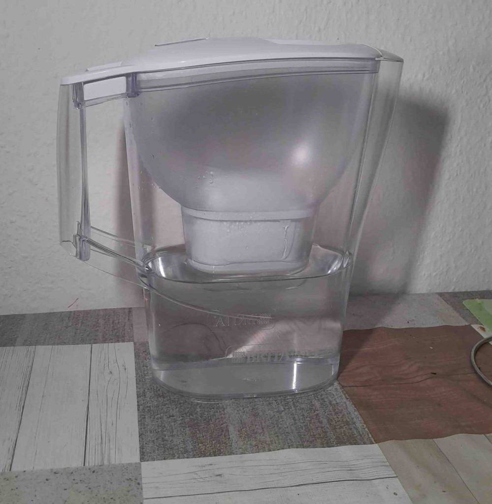 Im Test: ein Brita Wasserfilter mit transparentem Kunststoff-Gefäß, inklusive weißem Filter und Deckel, gefüllt mit Wasser steht auf einer weiß-braun karierten Tischdecke.