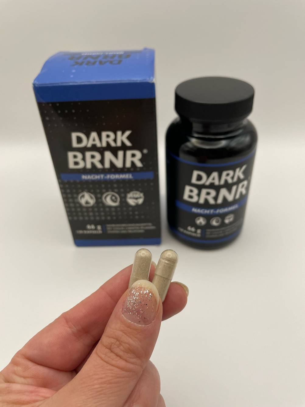 Im Test: Eine Hand hält zwei Kapseln Fatburner zwischen Daumen und Zeigfinger, im Hintergrund das Glas mit Verpackungskarton.