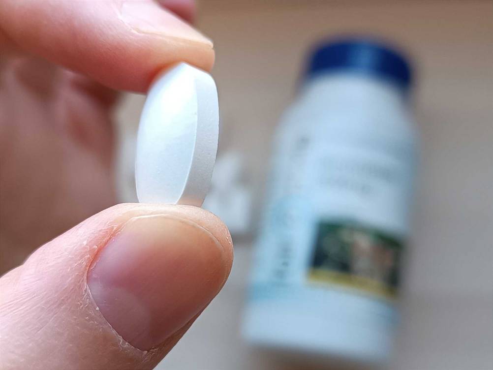 Eine Hand hält eine Tablette des getesteten Bromelain zwischen den Fingern, im Hintergrund die Packung.