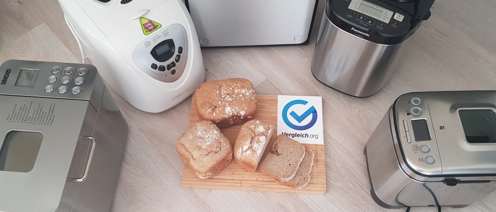Verschiedene Brotbackautomaten stehen im Kreis, in der Mitte ein aufgeschnittenes Brot.