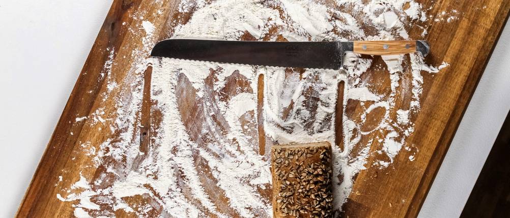 Brotmesser im Test: Auf einem bemehlten Holzbrett liegt ein Brotmesser und ein eckiger Brotlaib. Die Klinge ist mehr als doppelt so lang wie der Brotlaib breit ist.