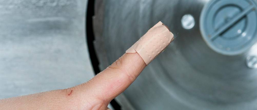 Finger mit Pflaster vor einer Messerscheibe einer Brotschneidemaschine manuell im Test.