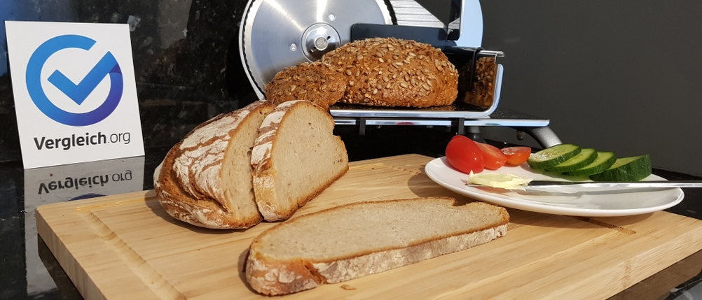 Brotschneidemaschine im Hintergrund, im Vordergrund geschnittenes Brot, sowie Gurken und Tomaten auf einem Teller