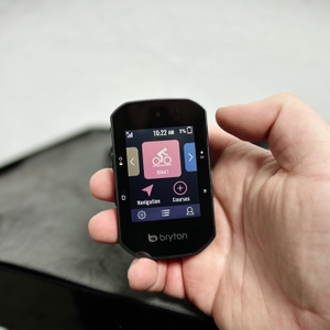 Ein Bryton Rider S500 wird mit der Menüansicht in der Hand gehalten. Das Display zeigt die Startseite mit verschiedenen Navigationsfunktionen.