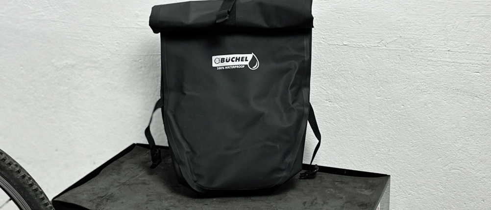 Die Büchel-81518001-Fahrradtasche steht frontal auf einem Schrank mit sichtbarem Logo. Der Rollverschluss ist nach vorne gerichtet.