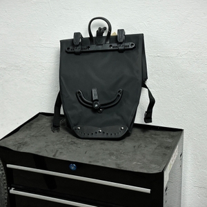 Die Rückseite der Büchel-81518001-Fahrradtasche mit zwei Befestigungshaken und einem verstellbaren unteren Haltebügel. Die Tasche steht aufrecht auf einem Werkzeugschrank.