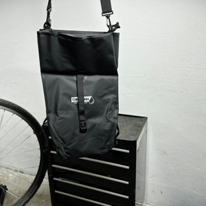 Die Büchel-81518001-Fahrradtasche wird an ihrem Schultergurt hängend getragen. Das Fahrrad steht im Hintergrund, die Tasche ist geschlossen.