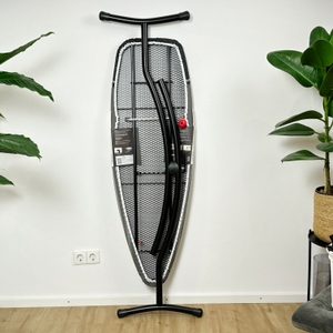 Das Brabantia-345647J-Bügelbrett steht zusammengeklappt an der Wand, sodass die Rückseite zu sehen ist. Die Rückseite der Verpackung zeigt Produktinfos.