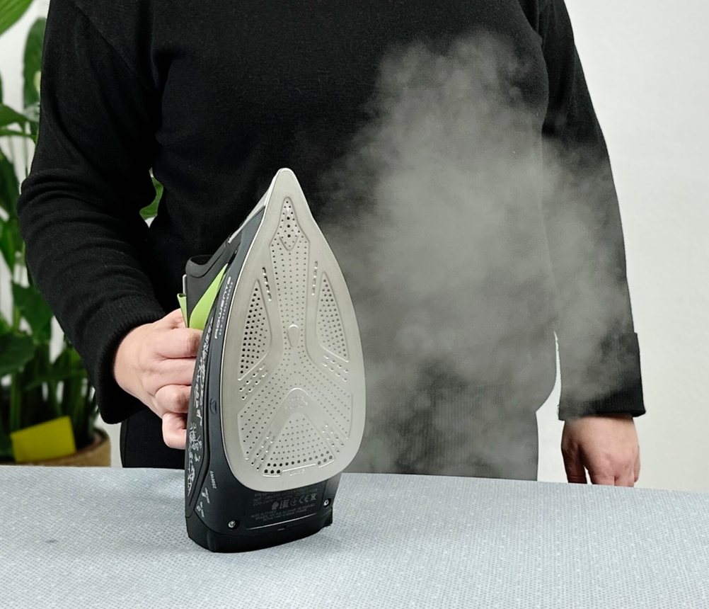 Eine Person in schwarzer Kleidung hält das Rowenta DW6330D1 Eco Intelligence Dampfbügeleisen senkrecht auf einem grauen Bügelbrett. Sichtbarer Dampf tritt aus der Bügelsohle aus.