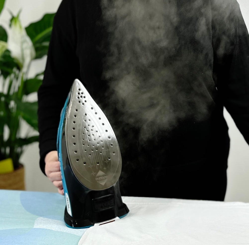 Eine Person in schwarzer Kleidung hält das Tefal FV9844 Dampfbügeleisen senkrecht auf einem Bügelbrett. Sichtbarer Dampf tritt aus der Bügelsohle aus, während ein weißes Hemd auf dem Bügelbrett liegt.