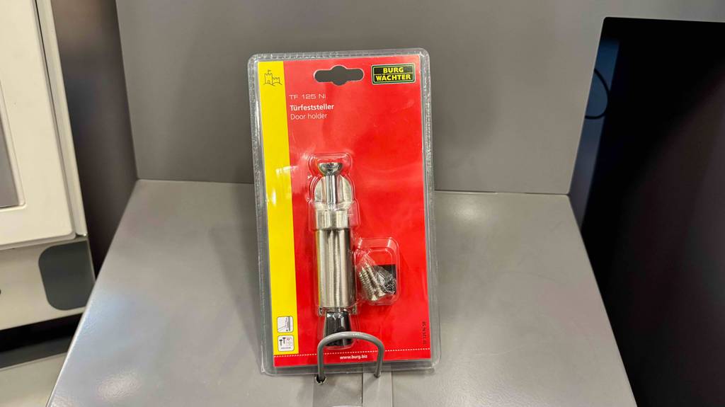 Im Test: Der Türfeststeller befindet sich eingeschweißt in einer transparenten Kunststoffverpackung inklusive roter Kartonbasis auf einem grauen Regal.