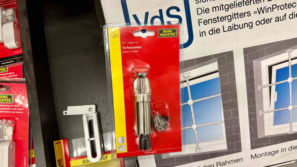 Getestet: Der Türfeststeller in der transparenten Kunststoffverpackung inklusive roter Kartonbasis befindet sich voer einem Plakat, auf dem Fenstergitter zu erkennen sind.