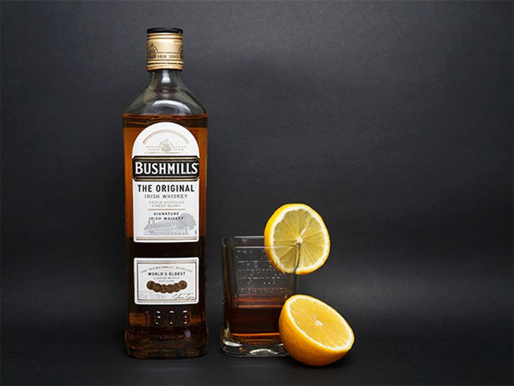 Bushmills im Test: Eine Flasche Bushmills The Original mit einem eingeschenkten Glas. Dazu eine Scheibe Orange. 