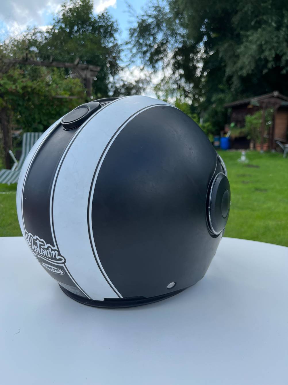 Ein schwarzer Retro-Jethelm mit weißen Streifen steht auf einem Tisch in einem Garten und ist von hinten zu sehen.