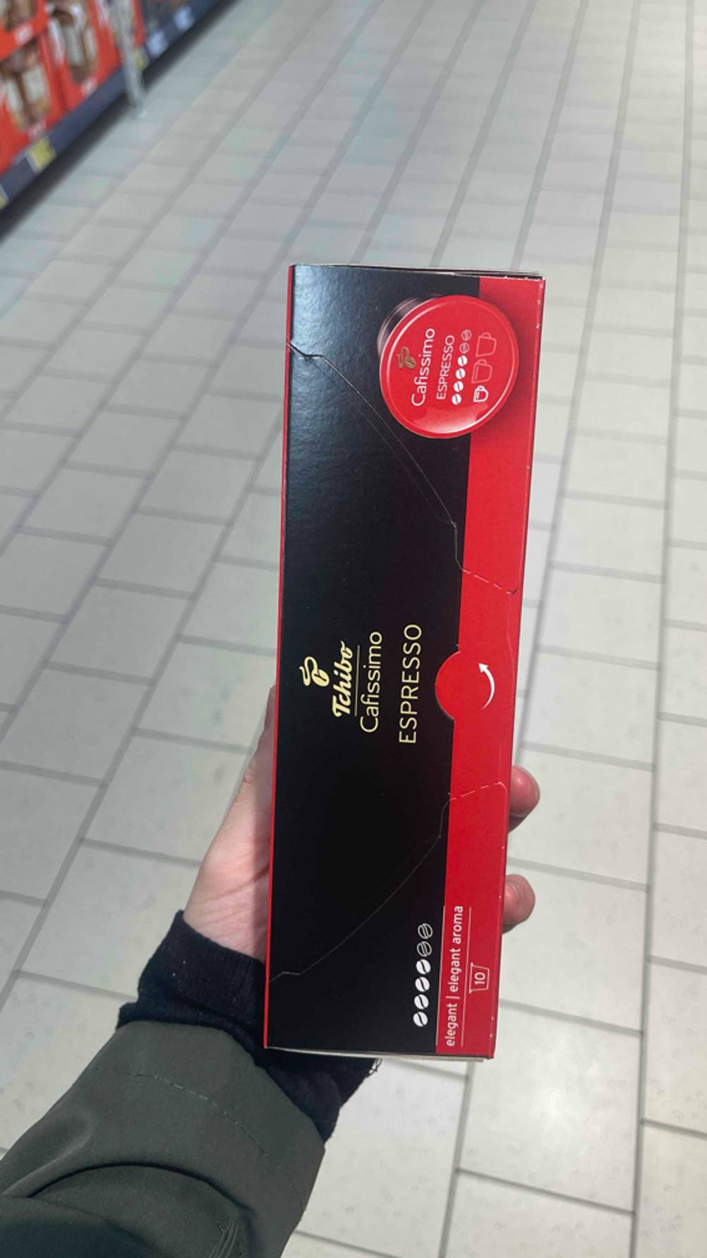 Cafissimo-Kapseln im Test: Eine Hand hält die schwarz-rote Packung mit 10 Stück über einem weißen Fliesenboden.