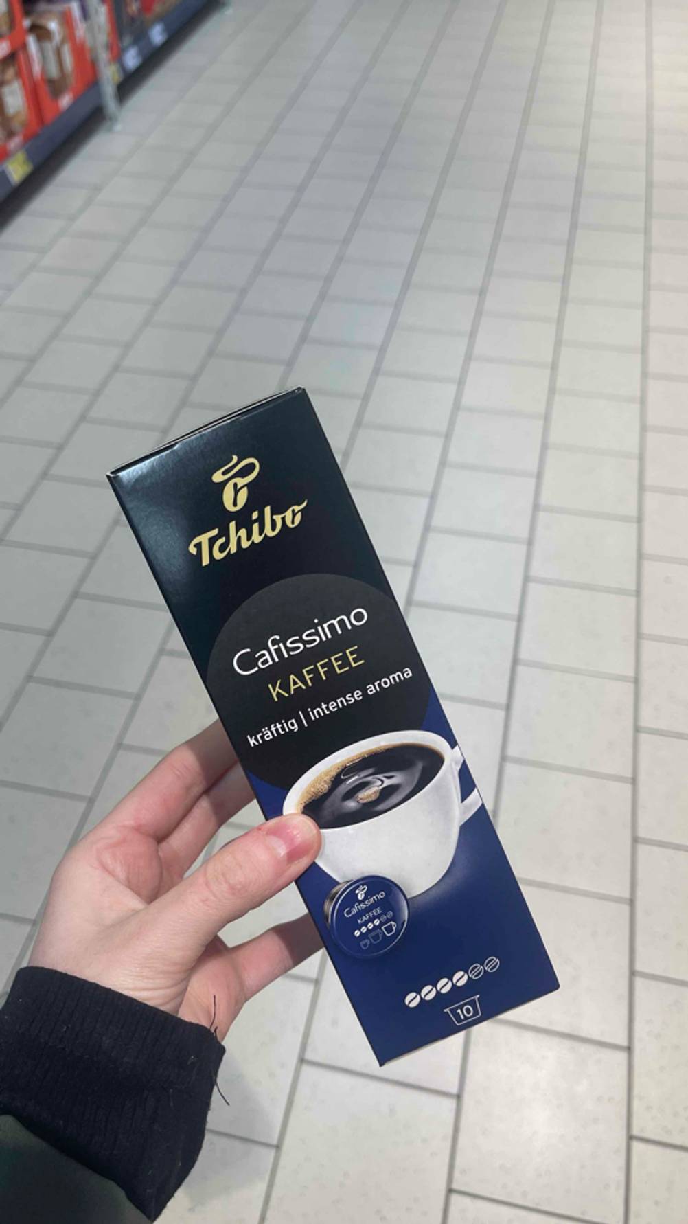 Eine Hand hält die schwarz-dunkelblaue Packung inklusive der getesteten Cafissimo-Kapseln „Kräftig“ über einem weißen Fliesenboden.