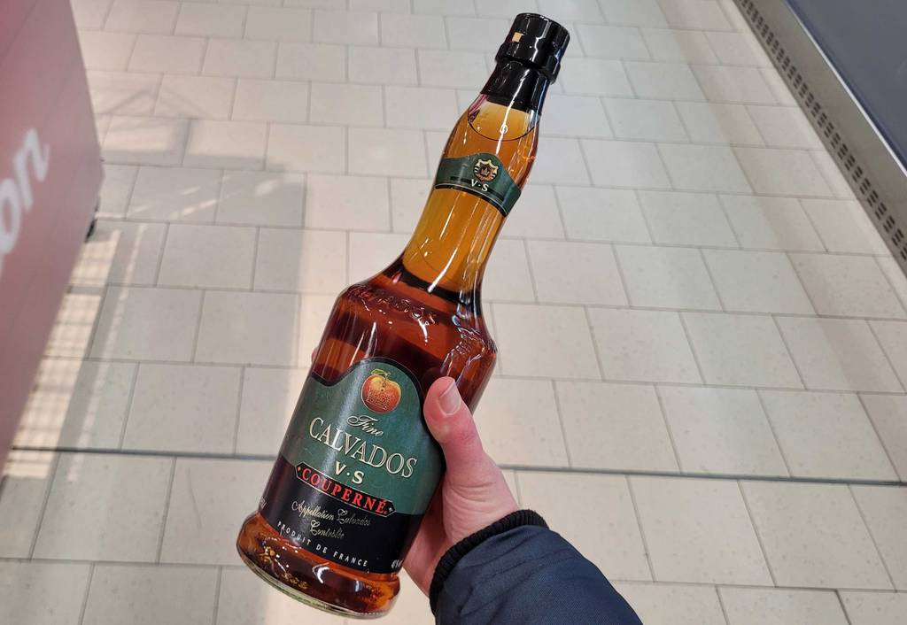 Hand hält eine transparente Glasflasche mit schwarzem versiegelten Verschluss eines Calvados Brandys im Test in einem Verkaufsbereich.