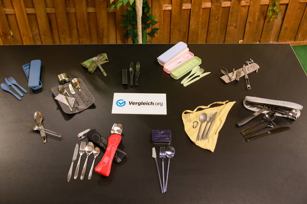 Camping-Besteck-Set-Test: Alle Camping-Besteck-Sets liegen mit einem Schild von Vergleich.org auf einem Tisch.