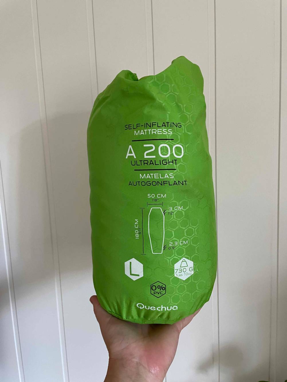 Eine Hand hält eine in einen Sack mit Logo verpackte Camping-Isomatte vor eine helle Wand.