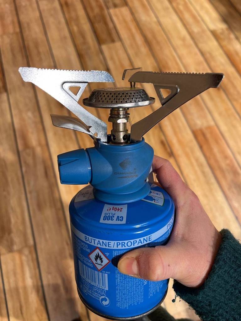 Hand hält einen Gaskocher im Test inklusive blauer Butan-Ventil-Kartusche über einen Holzboden.