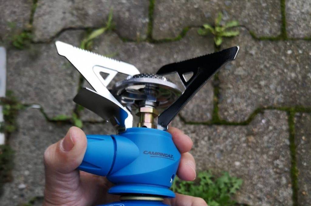 Gaskocher-Test: ein blauer Campingaz-Kompaktbrenner inklusive Kartusche in einer Hand vor grauen Pflastersteinen.
