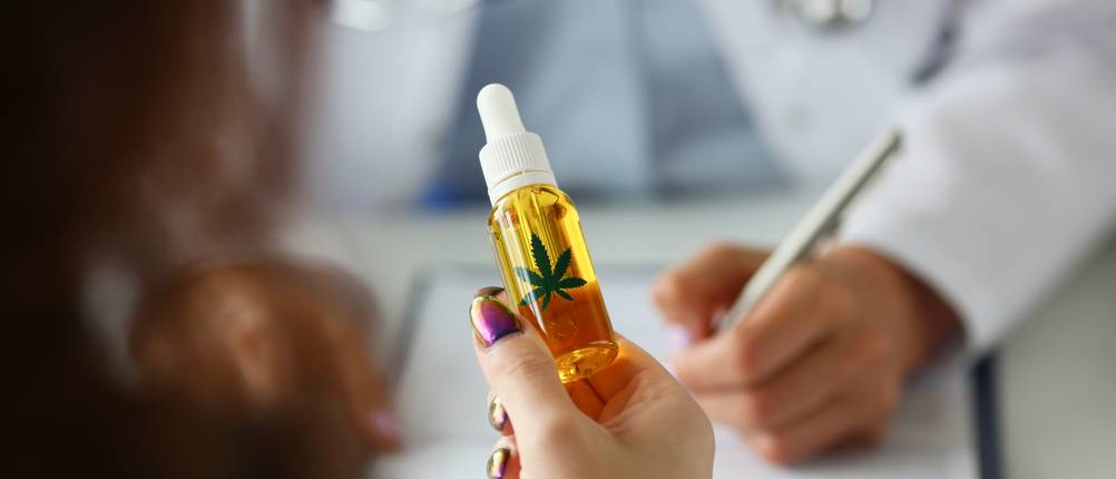 Cannabis-Rezepte im Test: Ein Person hält eine kleine Flasche mit Cannabis-Öl in der Hand, ihr gegenüber sitzt ein Arzt.