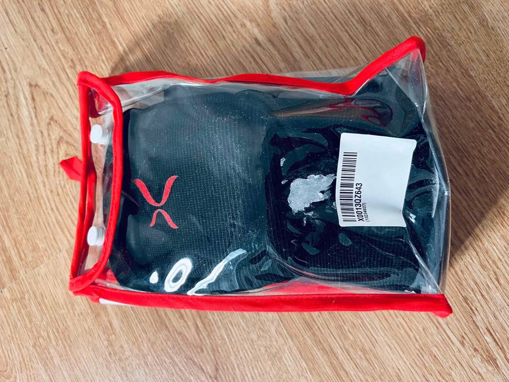 Schwarze Knieschoner im Test auf Holz, in wiederverschließbarer, transparenten Kunststoff-Originalverpackung mit rot abgesetzten Außennähten und weißen Druckknöpfen.