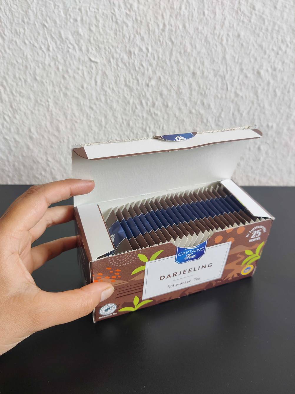 Eine Hand öffnet den Deckel einer Packung Darjeeling-Tee mit einzelnen Teebeuteln darin.