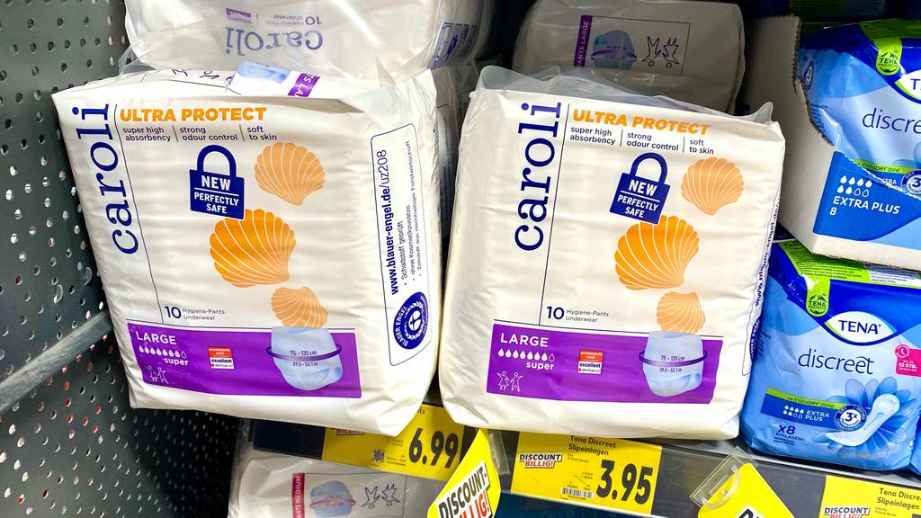 Im Test: Windeln für Erwachsene liegen in mehreren weißen Caroli Verpackungen neben Tena Einlagen in blauen Packungen auf einem Verkaufsregal.