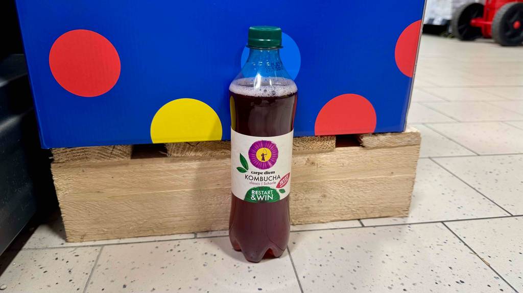 Kombucha-Test: Eine transparente Flasche mit roter Flüssigkeit steht vor einer Palette auf dem Boden.