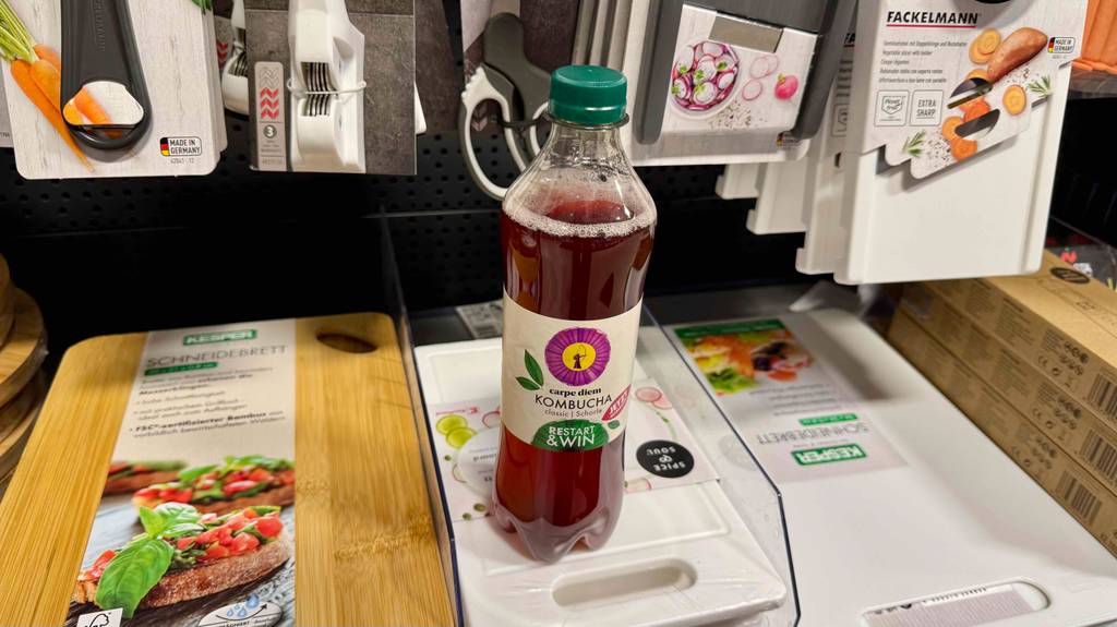Kombucha im Test: Eine transparente Flasche mit roter Flüssigkeit befindet sich auf einem Verkaufsregal mit Küchenutensilien.