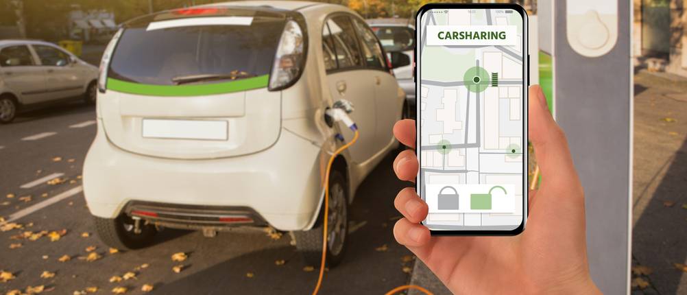 Carsharing-Test: Auf einem Smartphone ist die Carsharing-App geöffnet, während im Hintergrund ein Elektroauto lädt.