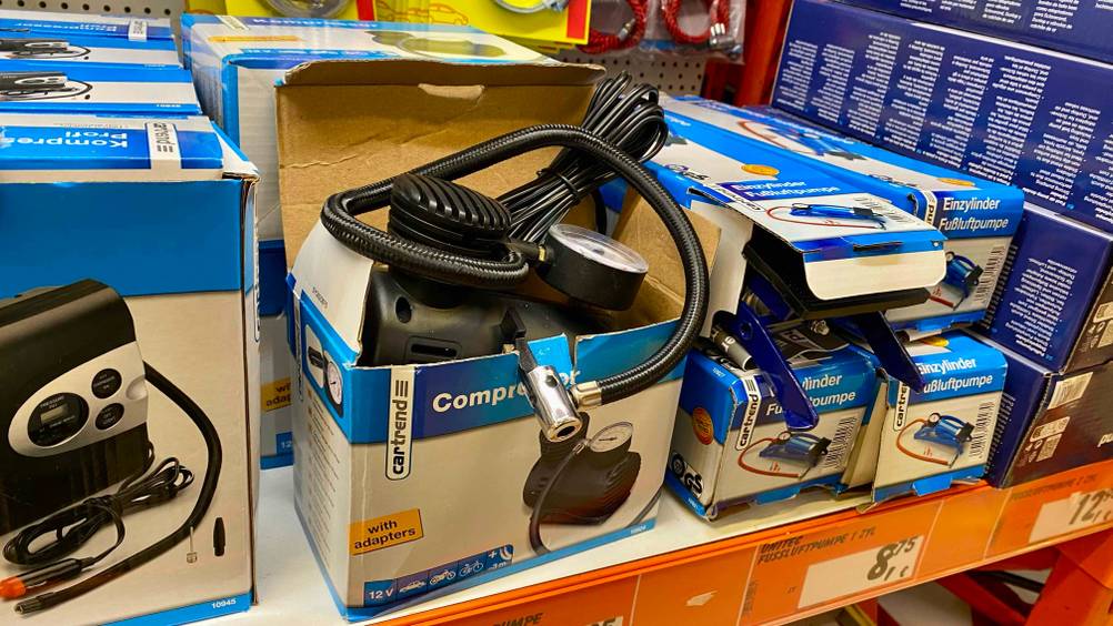 Ein getesteter 12V-Kompressor in seiner Originalverpackung mit geöffnetem Deckel neben anderen Produkten in einem Regal.