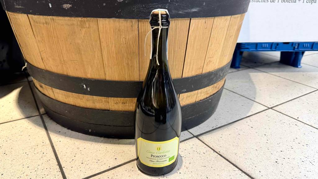 Bio-Wein im Test: Eine bauchige Prosecco-Flasche inklusive Korkenverschluss steht vor einem Holzfass.