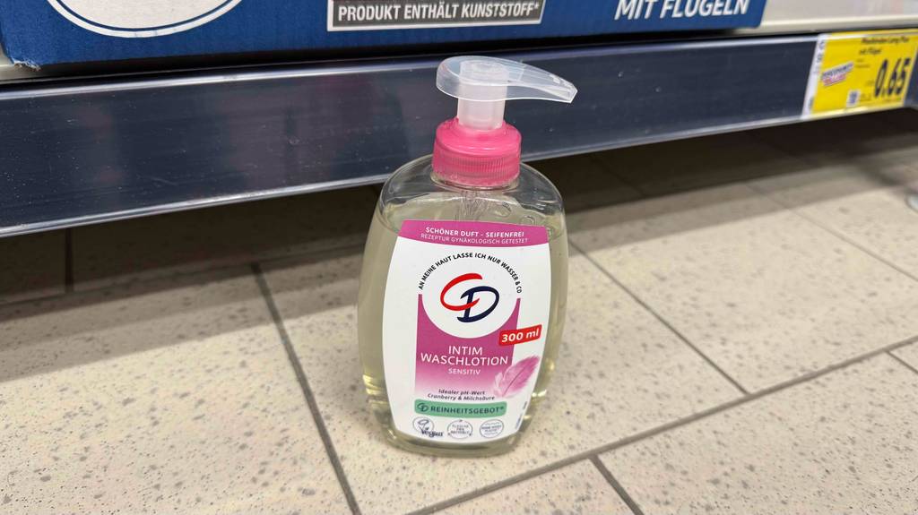 Eine Intimwaschlotion im Test inklusive eines transparenten 300-ml-Spenders steht vor einem Regal auf dem Boden.