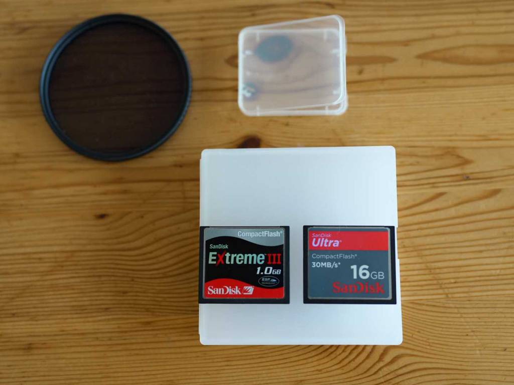 Zwei verschiedene CF-Karten im Test der Marke SanDisk liegen auf einer UV-Filter-Schutzhülle neben einem UV-Filter und einer CF-Kartenschutzhülle auf einer Holzfläche.