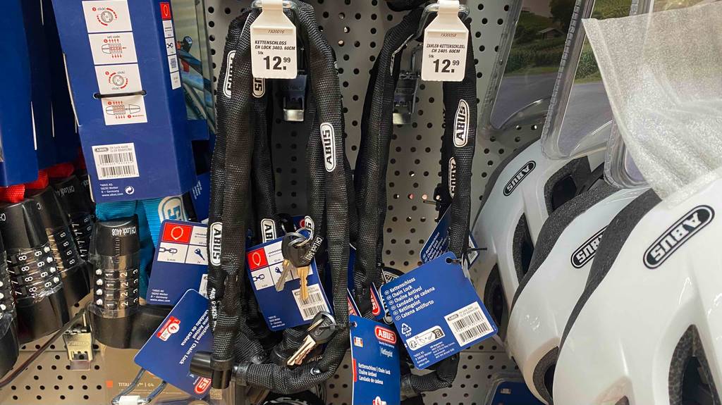 Mehrere mit schwarzem Gewebe überzogene ABUS-Kettenschlösser im Test hängen inklusive Schlüssel und blauem Pappticket an einem Verkaufsstand mit ABUS Fahrradzubehör.