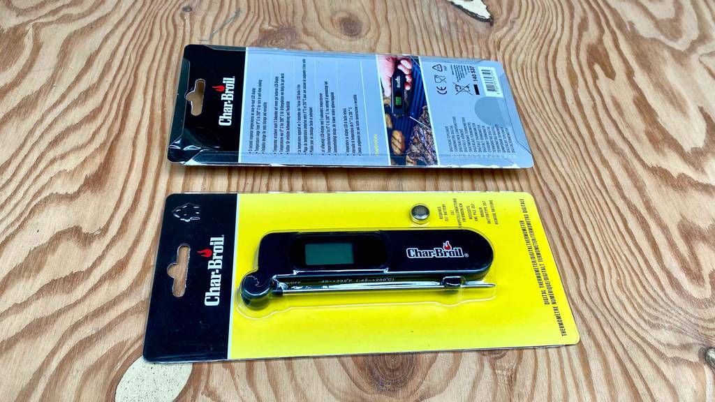 Zwei Verpackungen eines digitalen Char-Broil Grillthermometers im Test, mit gelber Vorderseite inklusive eingeschweißtem Thermometer und Knopfbatterie, sowie hellblauer Rückseite mit Produktinformationen, liegen auf einer Holzfläche.
