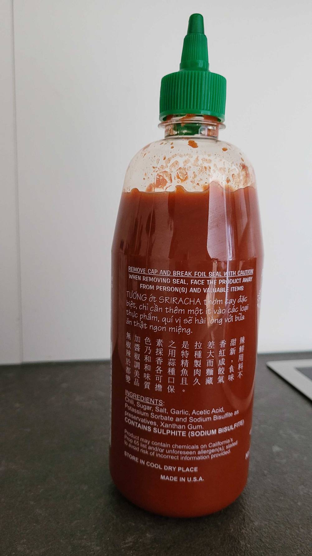 Rückseite einer Flasche der getesteten Chili-Sauce auf einer dunklen Fläche vor einer hellen Wand.