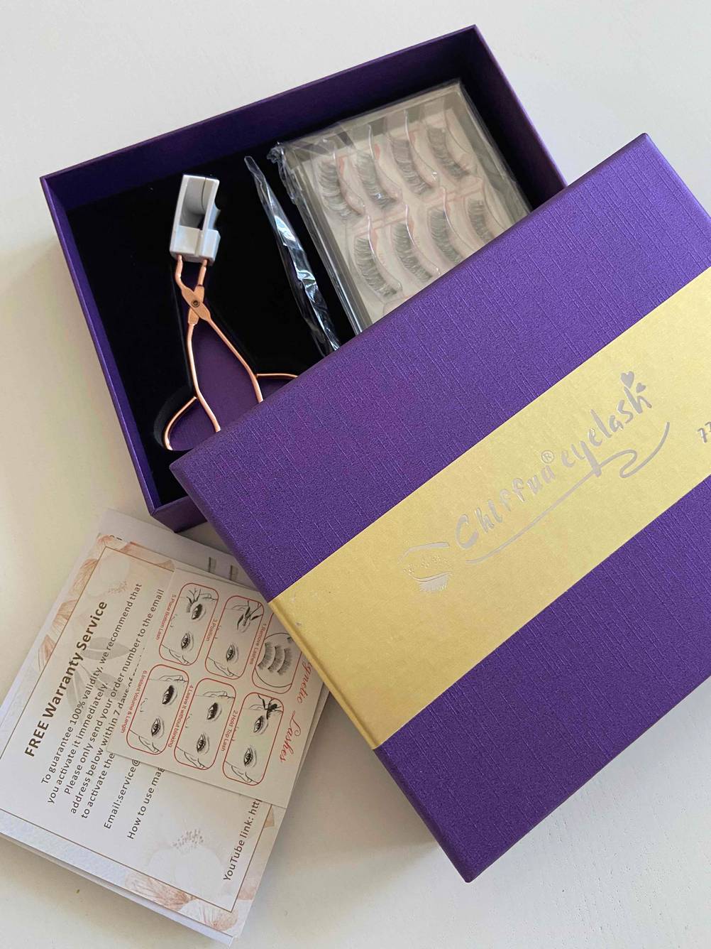 Original verpacktes Zubehör in gold- und lilafarbener Geschenk-Schachtel von magnetischen Wimpern im Test auf heller Fläche.