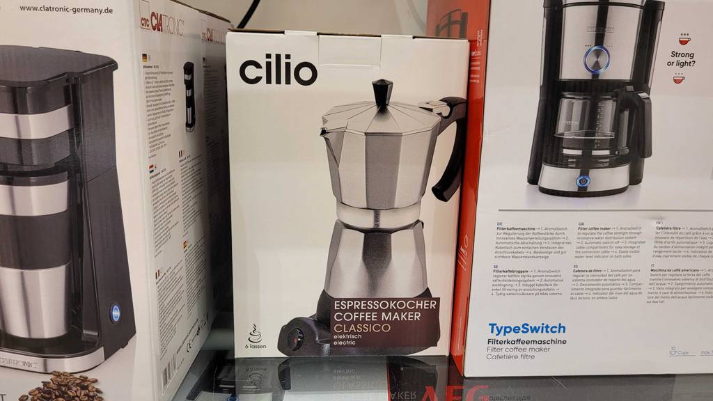 Ein Espressokocher elektrisch in Originalverpackung steht zum testen in einem Regal neben anderen Geräten.