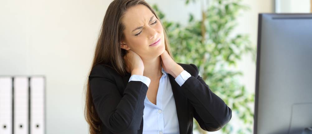 Businesswoman suffering neck pain sitting in a chair while working with a desktop computer in her workplace at office
