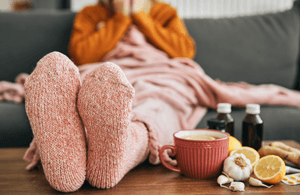 Cistus-Kapseln-Test: Eine Frau sitzt mit dicken Socken auf einer Couch neben einem Glas Tee, Knoblauch und Tabletten. 