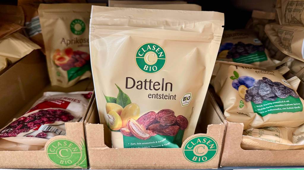 In einem Verkaufsregal steht eine wiederverschließbare Tüte mit entsteinten getesteten Bio-Datteln zwischen anderen Trockenfrüchte-Verpackungen.
