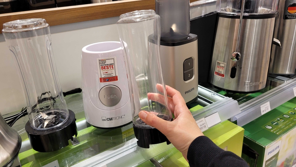 Smoothie-Maker im Test: Eine Hand hält den Mixbecher eines Smoothie-Makers vor ein Regal mit Produkten.
