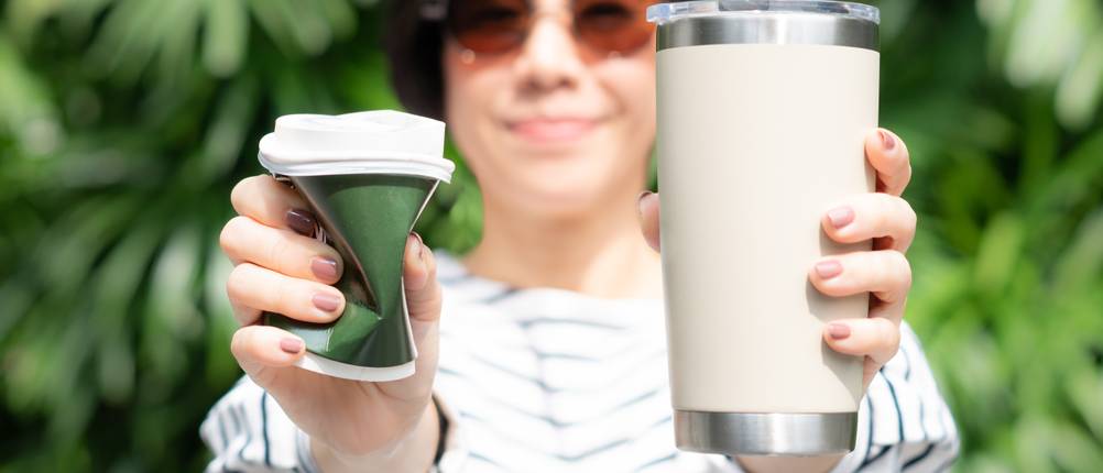 Coffee-to-go-Becher-Test: Eine Frau, die einen Coffee-to-go-Becher in beiden Händen hält. Einer ist ein Einweg-Papierbecher mit Plastikdeckel, der andere ein wiederverwendbarer Edelstahlbecher. Kein Strohhalm und das Zero-Waste-Konzept.
