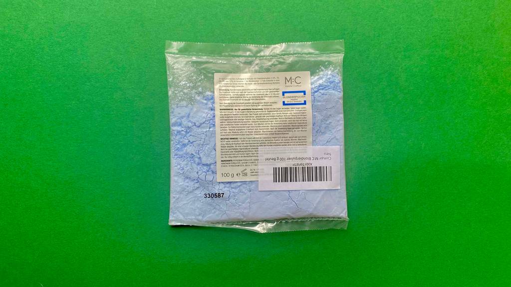 Vor einem grünen Hintergrund ist eine bläuliches Blondierpulver im Test in einer transparenten 100-g-Kunststofftüte.